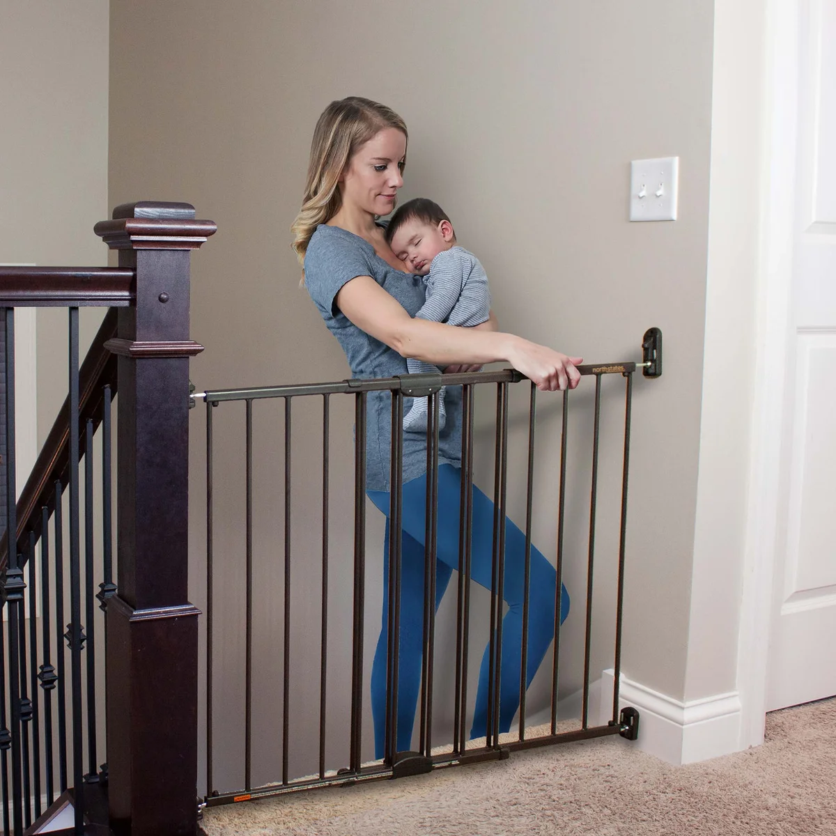 Mother holding sleeping baby while operating the Toddleroo Easy Swing & Lock gate one-handed at top of stairs