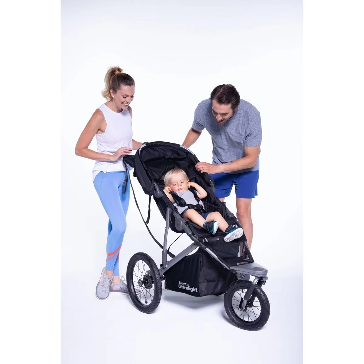 Parents loading sleeping baby into the Joovy Zoom 360 Ultralight stroller against white studio background