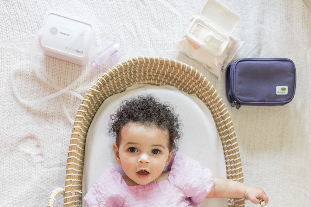 Overhead flatlay of the Dr. Noze Best NozeBot unit, tubing, nosepiece, wipes, and navy travel case beside a baby
