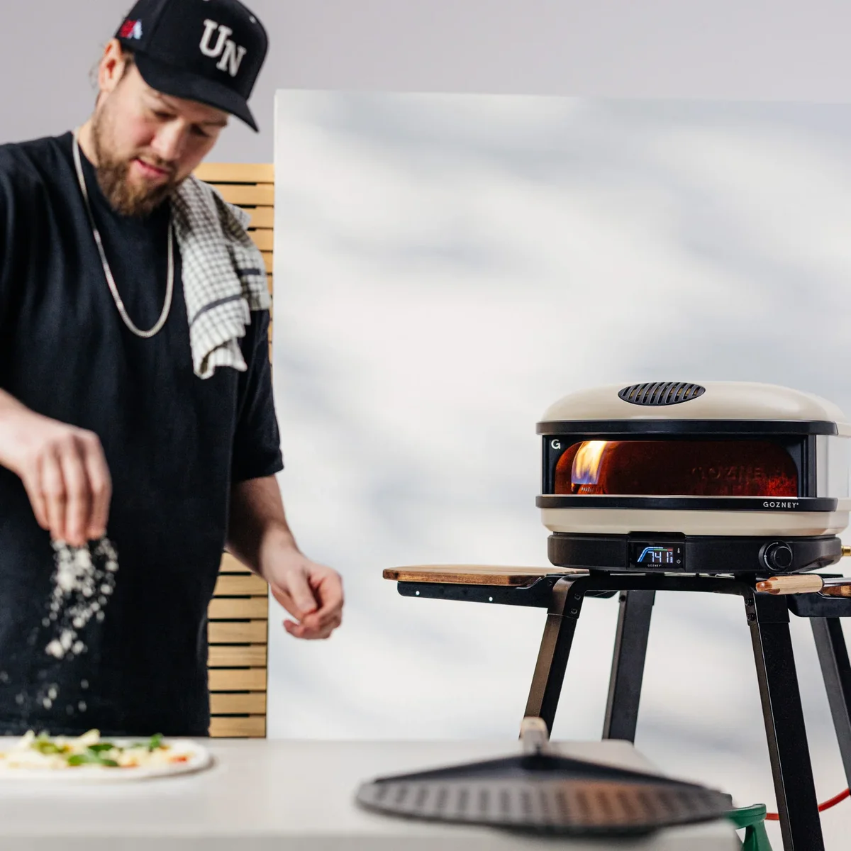 Gozney Arc XL pizza oven with visible flame in use outdoors while a cook prepares pizza dough in the background