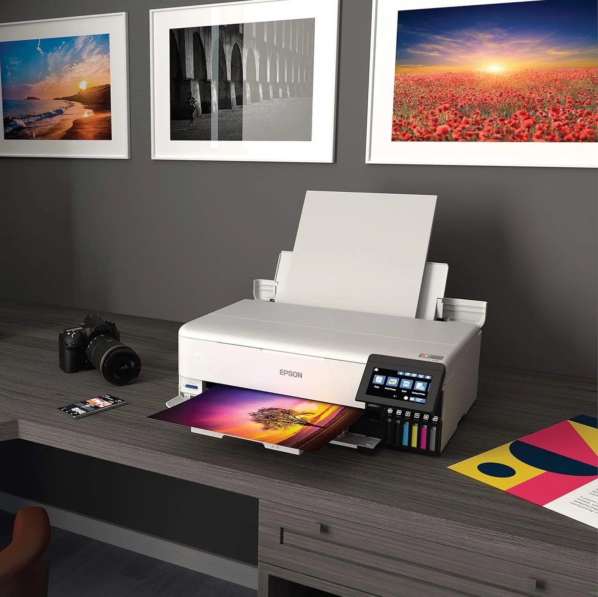 Epson EcoTank ET-8550 in use on a desk with a DSLR camera and framed prints displayed on the wall behind