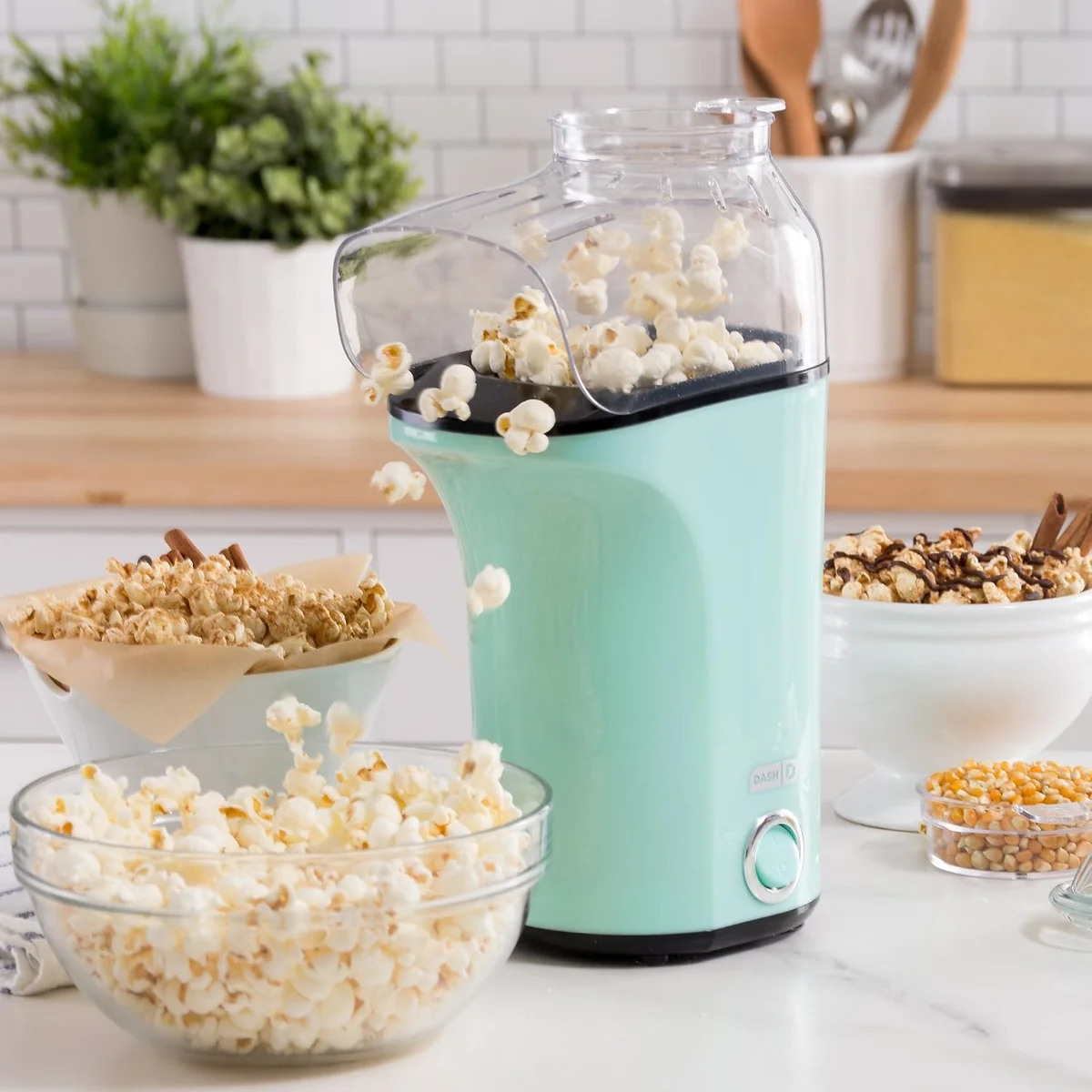 Dash Fresh Pop aqua popcorn maker on a kitchen counter with bowls of popcorn and kernels