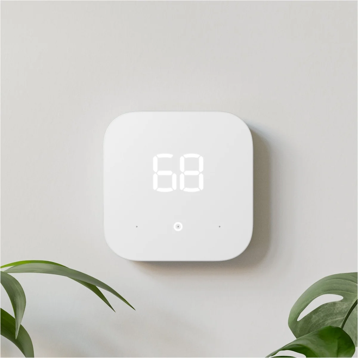 Amazon Smart Thermostat mounted on a light gray wall between two potted plant leaves, displaying 68 degrees