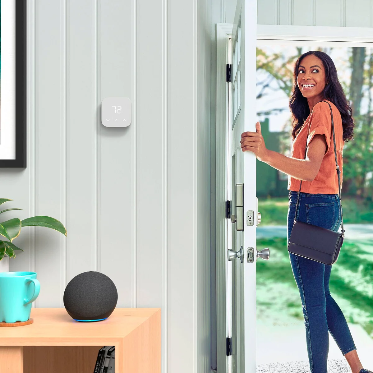 Amazon Smart Thermostat installed in a hallway showing 72 degrees, with an Echo Dot speaker nearby as a woman leaves through the front door