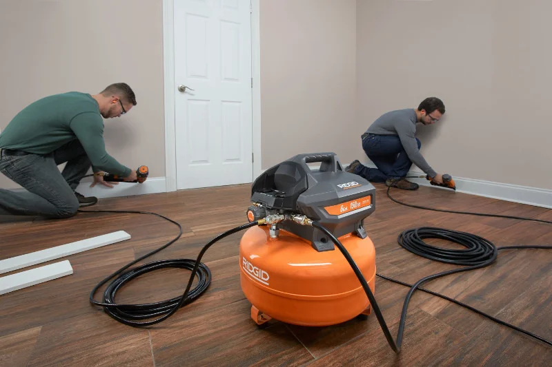 Two workers using the RIDGID 6-gallon pancake compressor to install trim and flooring, with air hoses running to pneumatic nailers