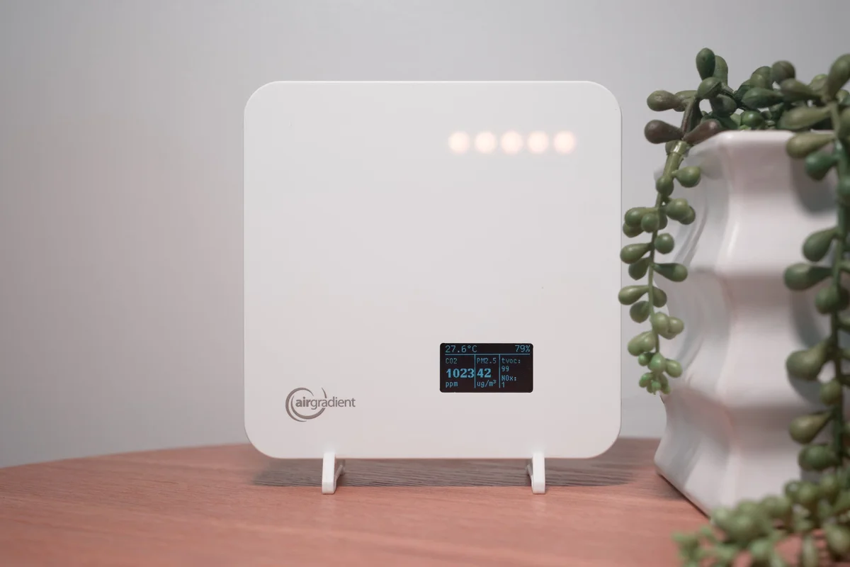 AirGradient One monitor in use on a wooden shelf beside a plant, OLED display showing live air quality readings