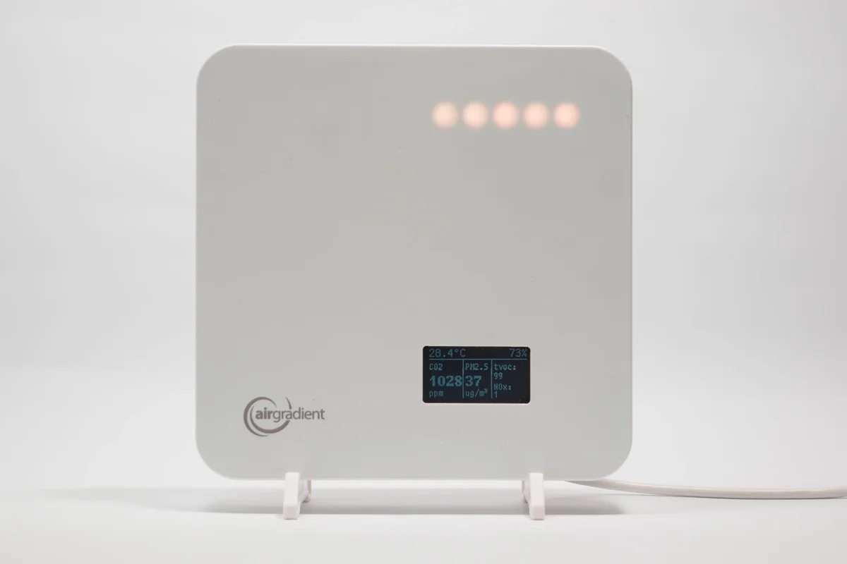 AirGradient One indoor air quality monitor front view, white enclosure with RGB LED bar and OLED display showing CO2, PM2.5, TVOC and NOx readings