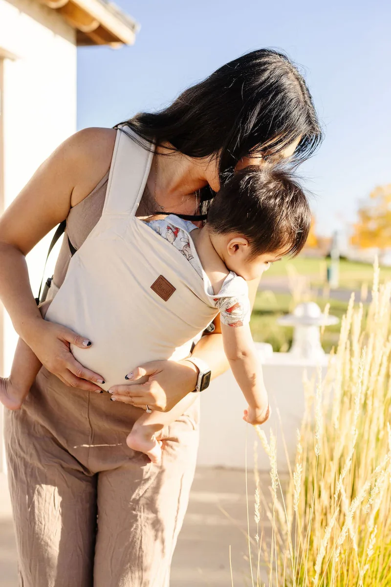 Happy Baby Original carrier in almond worn outdoors as mother leans over to let baby explore tall grass