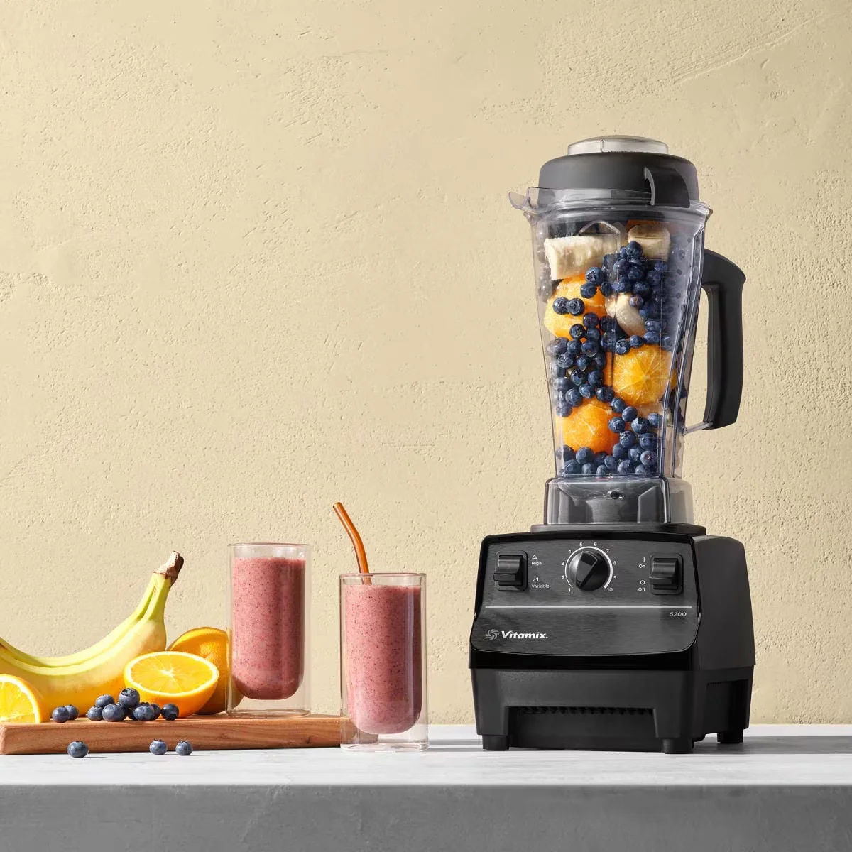 Vitamix 5200 on a kitchen counter with blueberries and mango in the pitcher, pink smoothie glasses, bananas, and sliced oranges