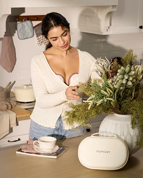 Momcozy M6 breast pump worn hands-free while woman arranges flowers in kitchen