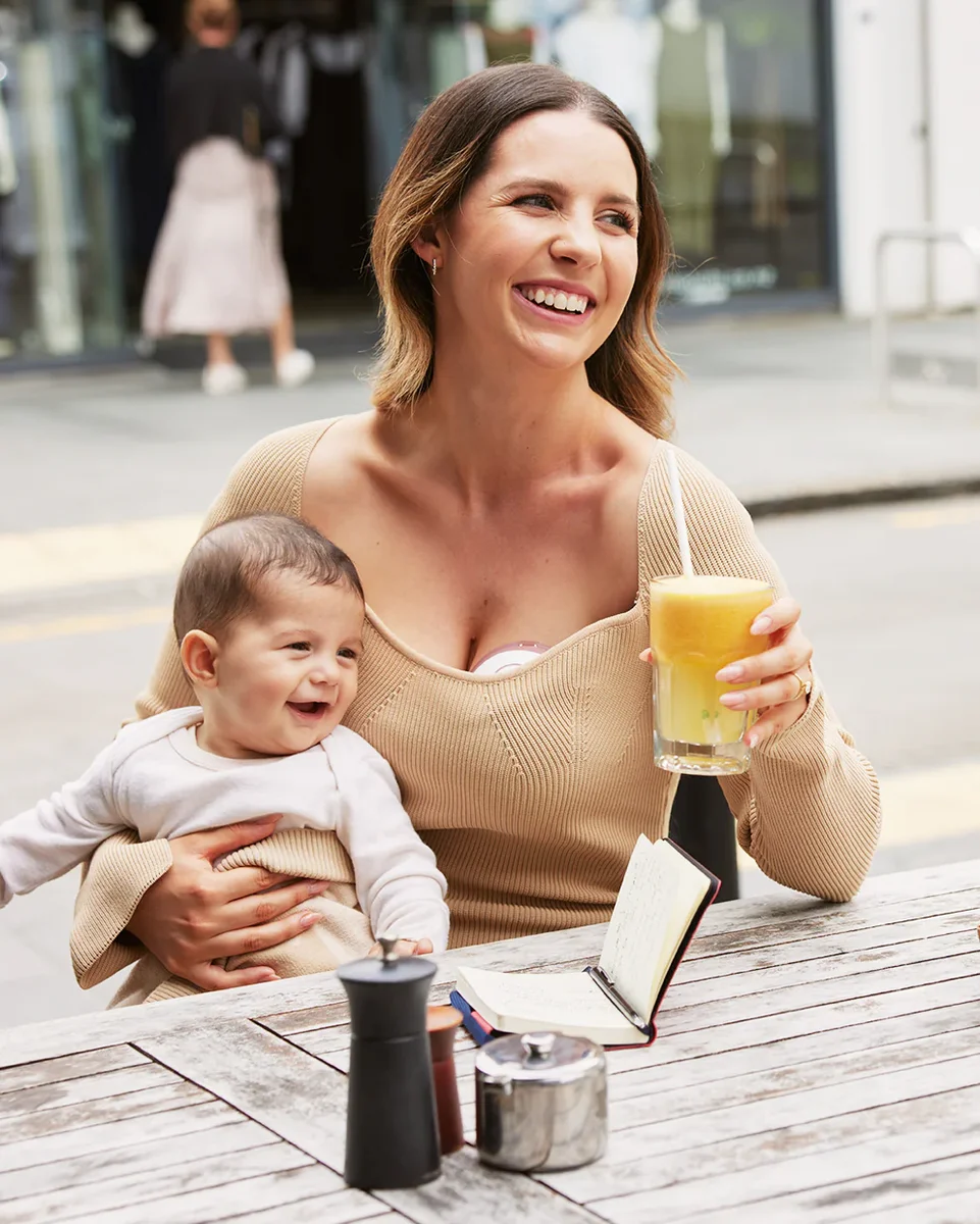 Mother using Momcozy M6 breast pump hands-free while holding baby at outdoor cafe