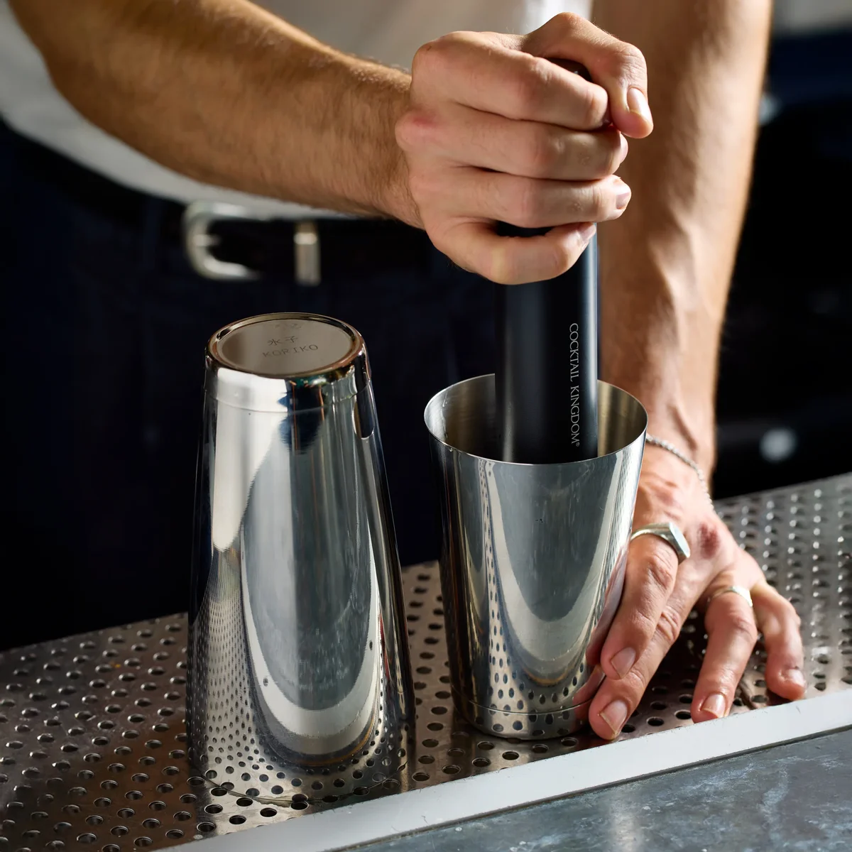 Koriko tins on a bar mat showing the stamped weighted base while a drink is muddled in the small tin