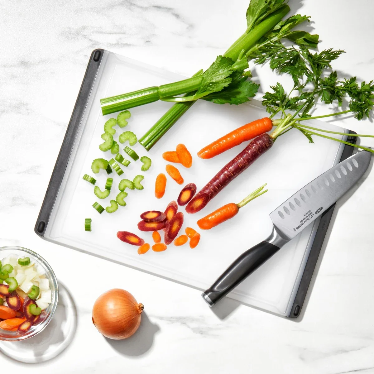 OXO Good Grips cutting board on a marble countertop with a chef's knife and sliced carrots and celery, showing the board's black rubber edge and feet