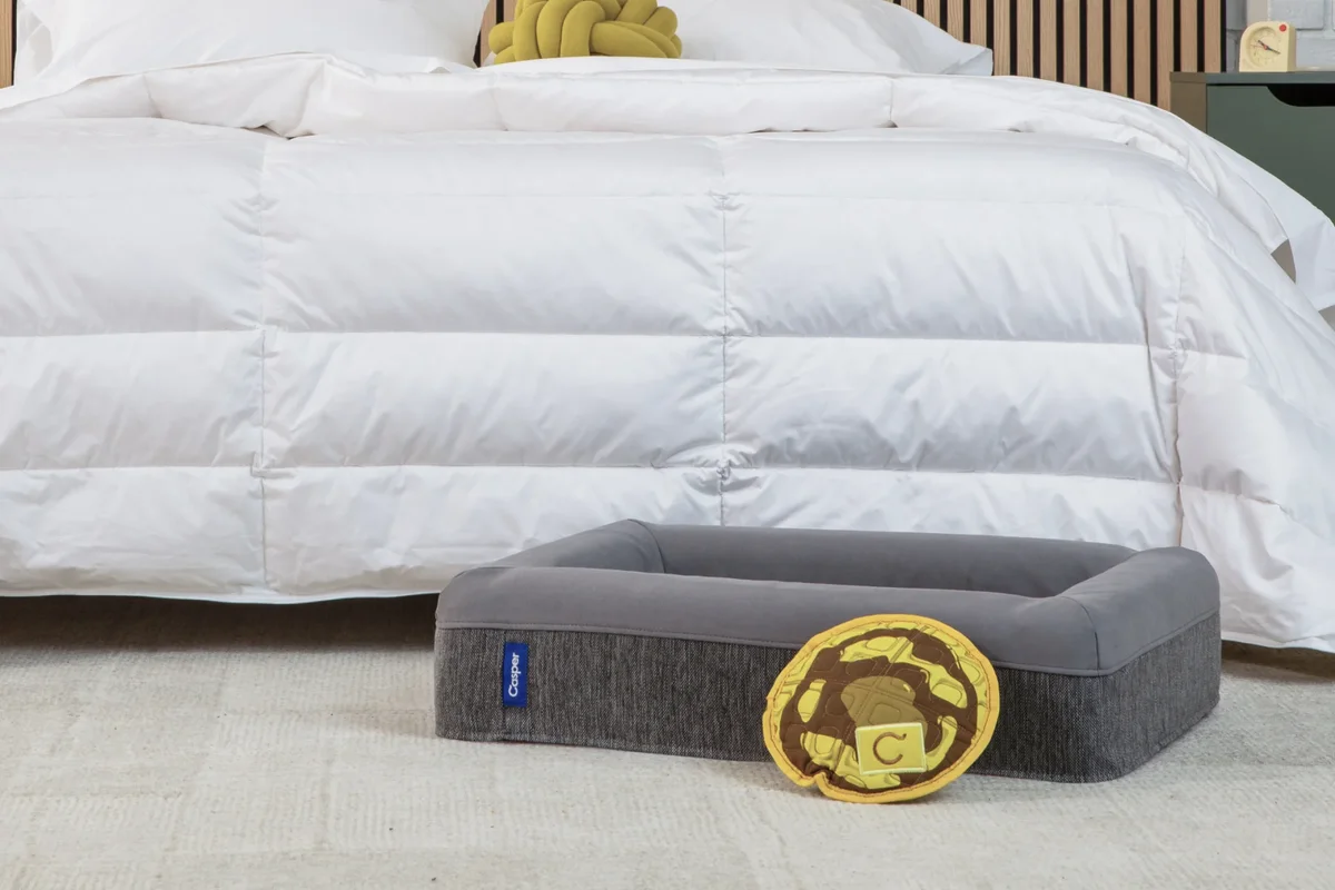 Casper Dog Bed in gray at the foot of a bed with the included waffle-shaped toy, lifestyle bedroom setting
