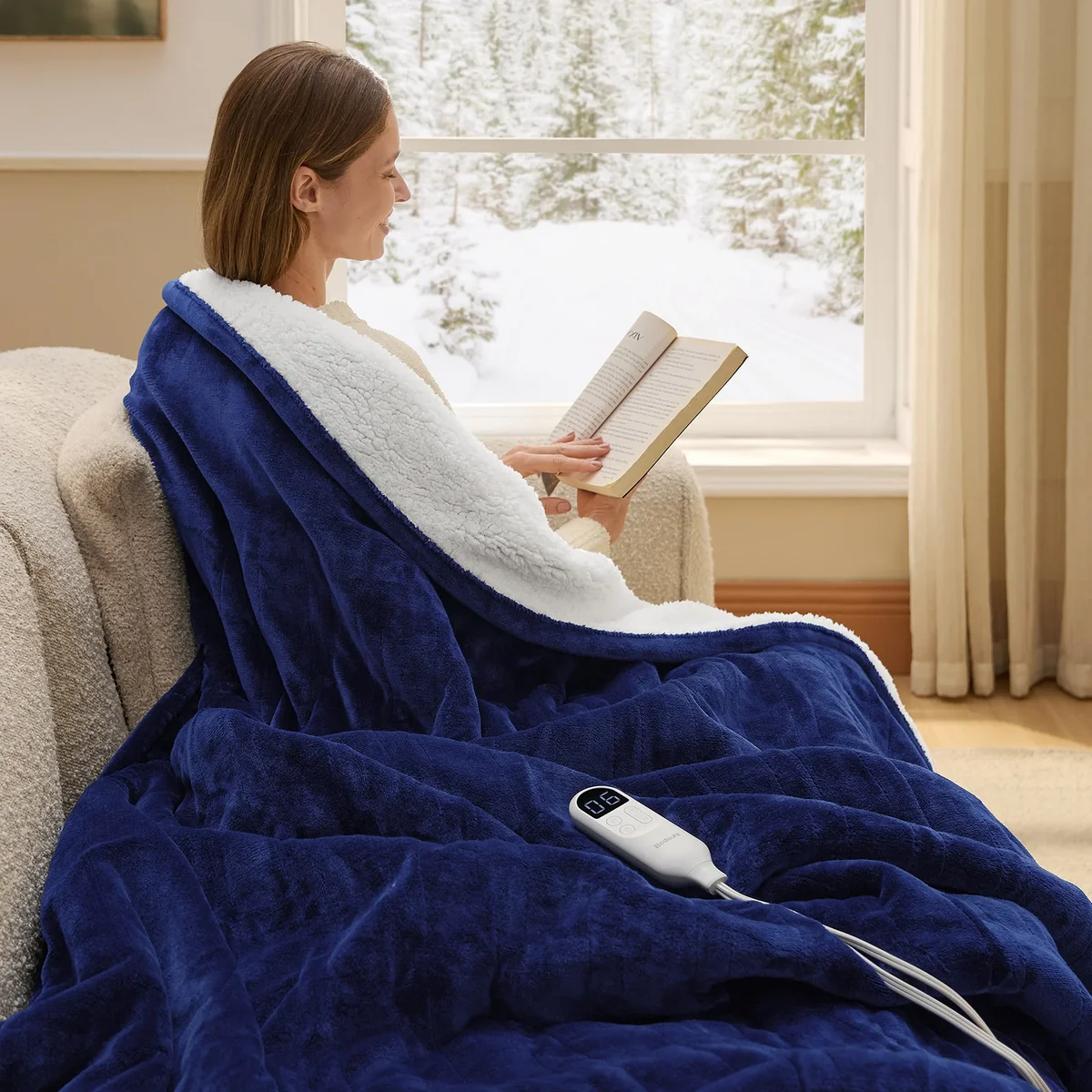 Woman reading while wrapped in the Bedsure GentleSoft heated blanket by a snowy window