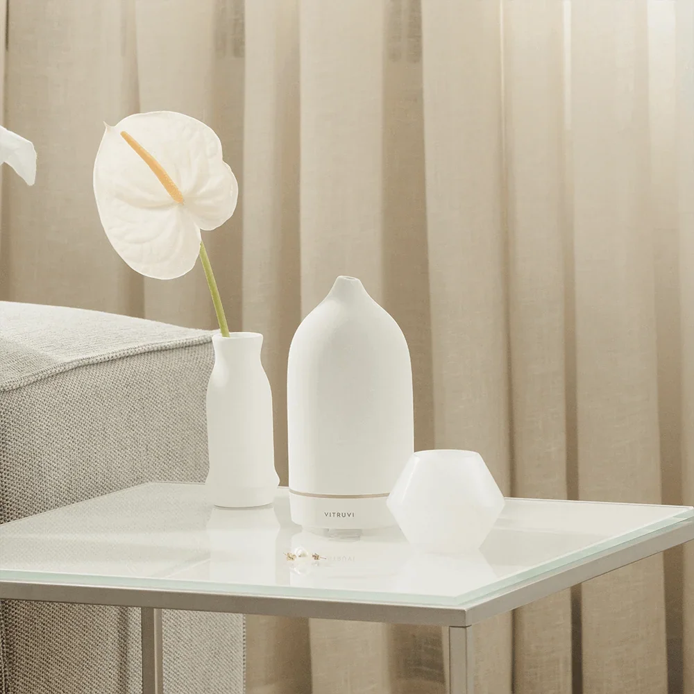 White Vitruvi Stone Diffuser styled on a glass side table beside a sofa, with a white anthurium in a vase and sheer linen curtains behind