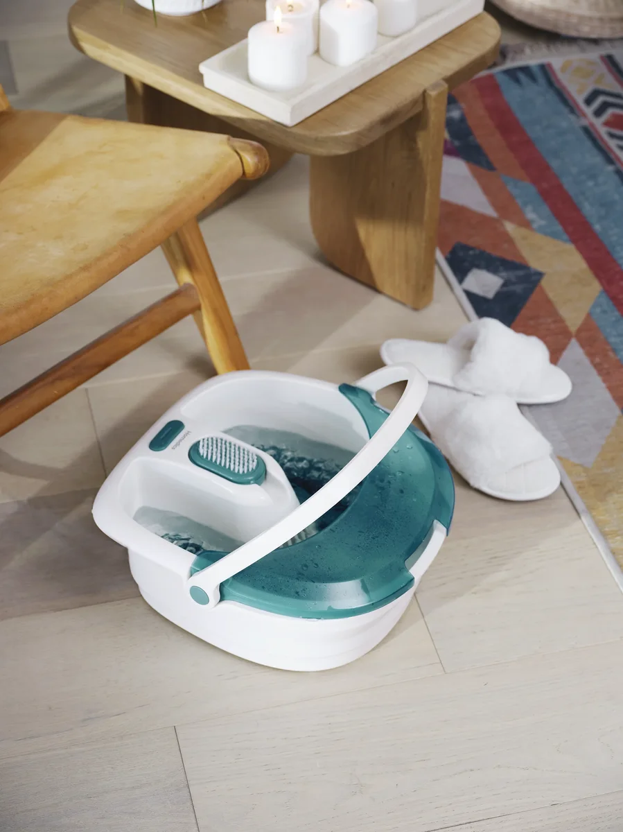 HoMedics Bubble Spa Elite Footbath on a wooden floor with its carry handle raised, beside slippers and a candlelit side table