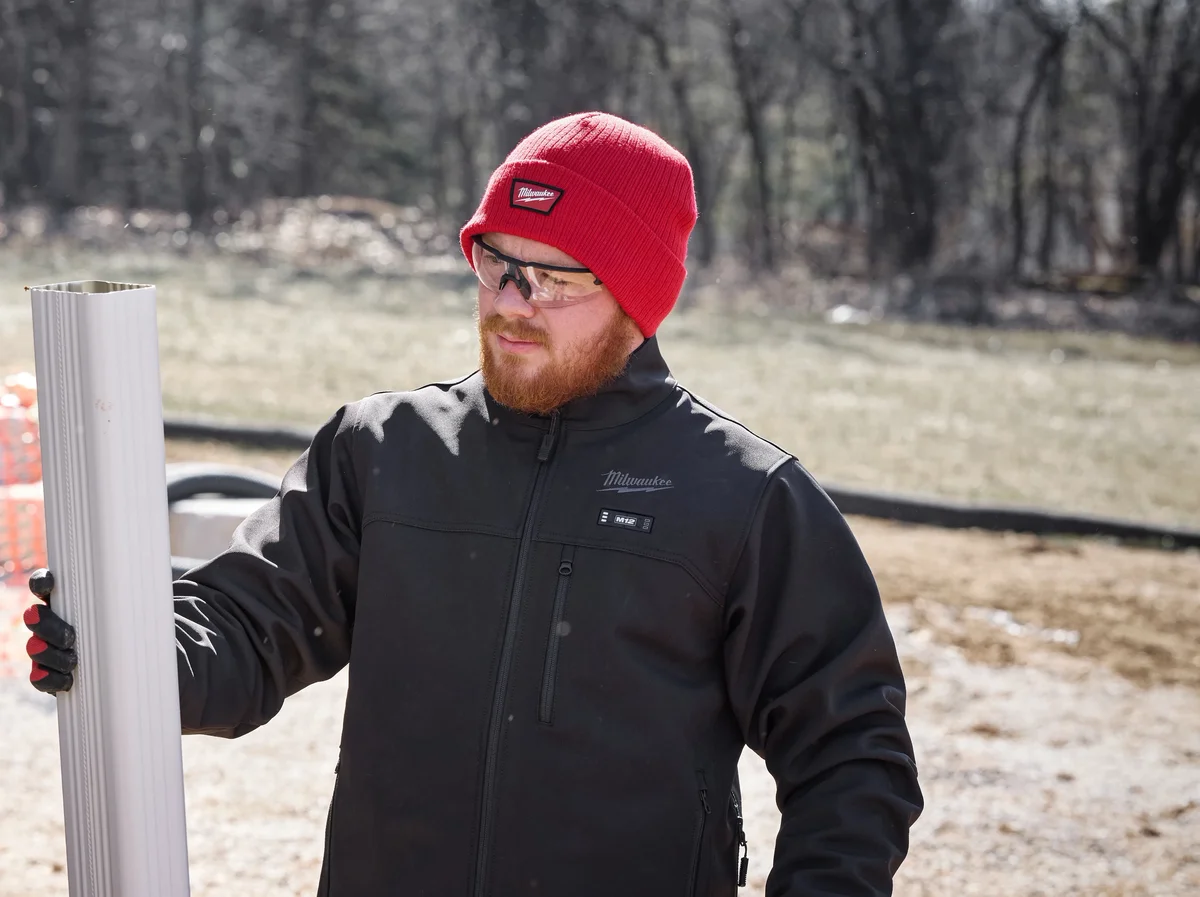 Worker in the black Milwaukee M12 Heated ToughShell Jacket carrying a length of downspout on a winter job site, with the M12 heat-control button on the chest