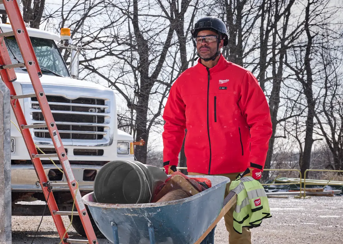 Worker in the red Milwaukee M12 Heated ToughShell Jacket pushing a wheelbarrow past a work truck on a snowy morning