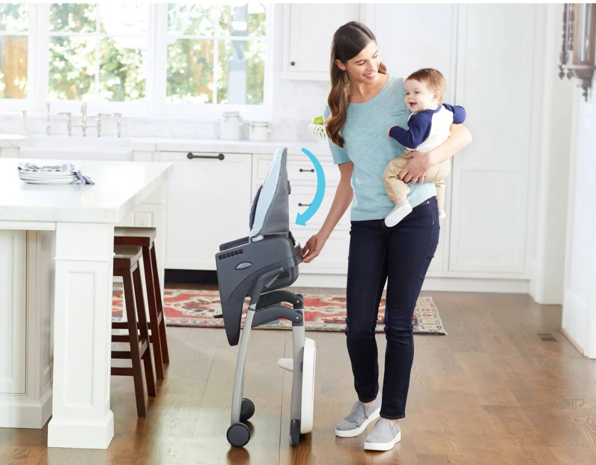 Graco DuoDiner DLX folded for storage — parent holding baby while folding the chair one-handed