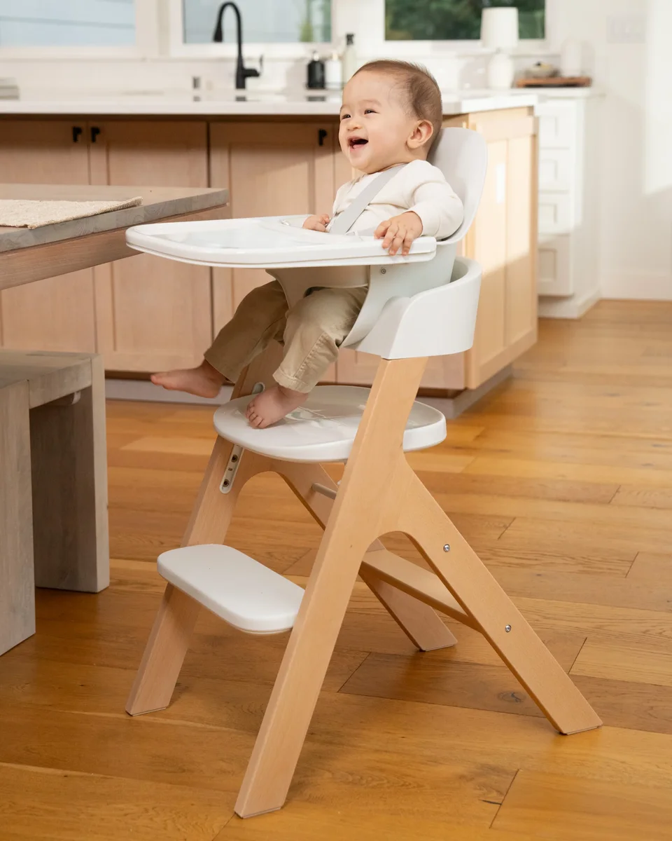 Baby sitting and smiling in Mockingbird High Chair at kitchen table, showing compact footprint