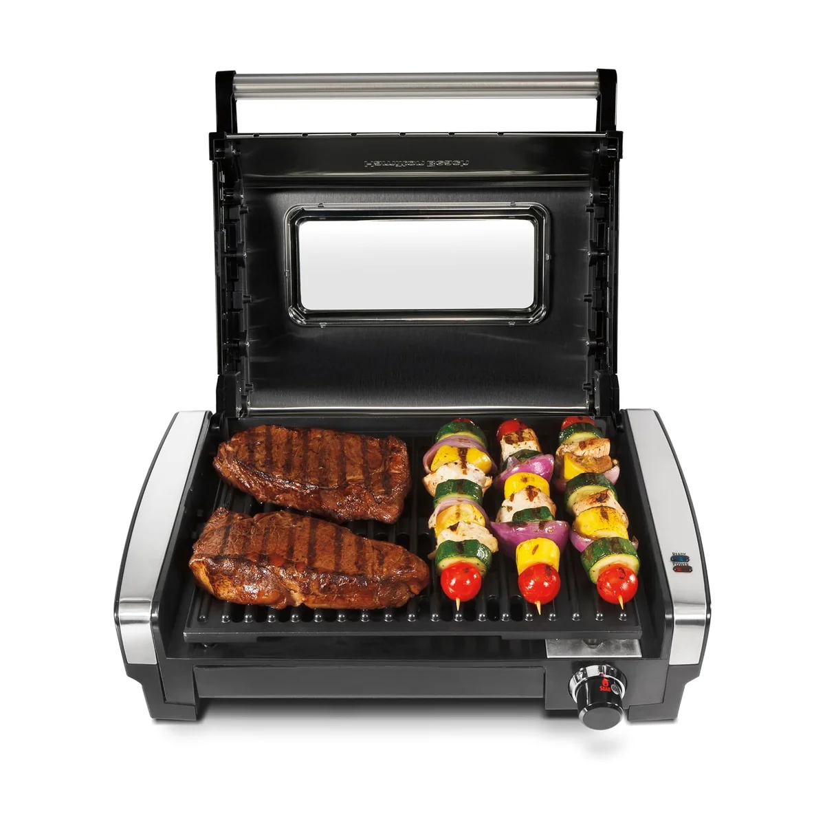 Hamilton Beach 25361 Searing Grill from the front with the hinged lid raised to show the viewing window, and steaks and vegetable kebabs on the grill plate