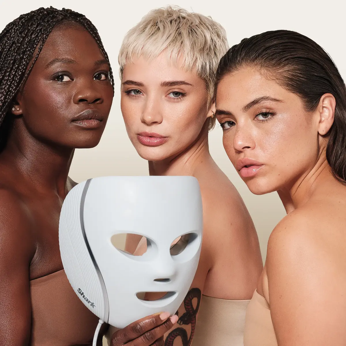 Three women posing with the Shark CryoGlow LED Face Mask held in front of them, showing the mask's ribbed side panel and silver trim
