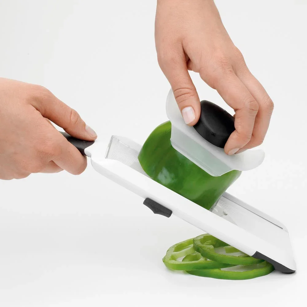 OXO Good Grips Hand-Held Mandoline Slicer in use, one hand gripping the handle while the other guides a green bell pepper with the food holder, rings falling below and the side thickness slider visible