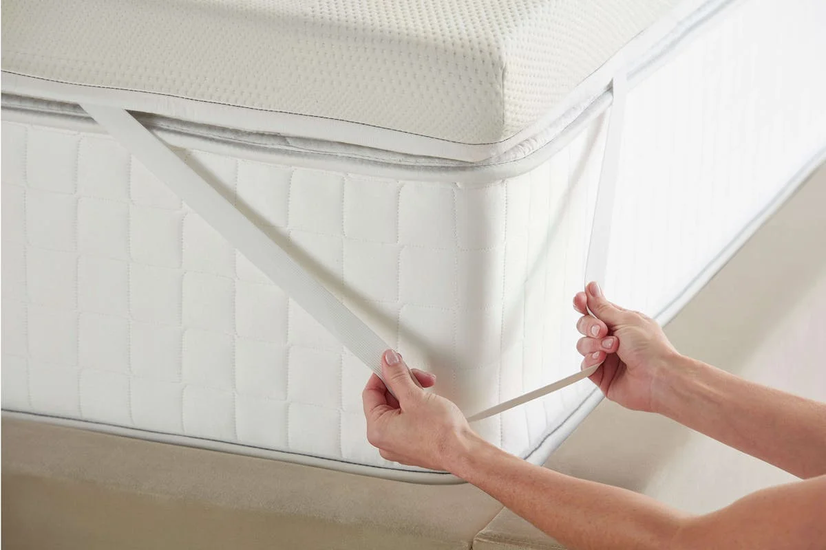 Close-up of hands attaching the elastic corner strap of the Saatva Graphite Memory Foam Mattress Topper to a mattress