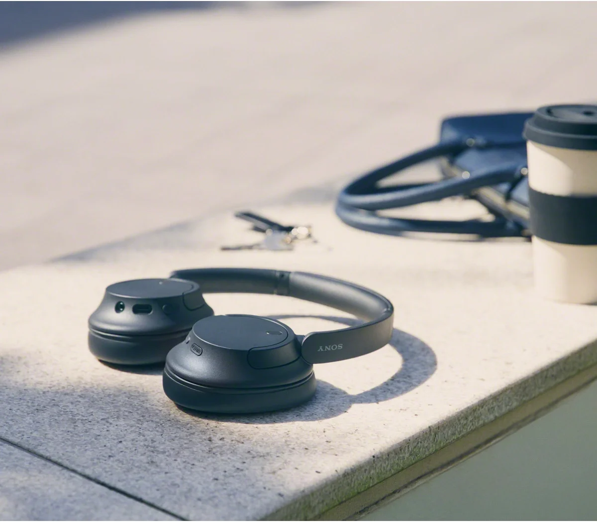 Sony WH-CH720N in Black resting on a sunlit stone ledge beside keys, a bag and a coffee cup