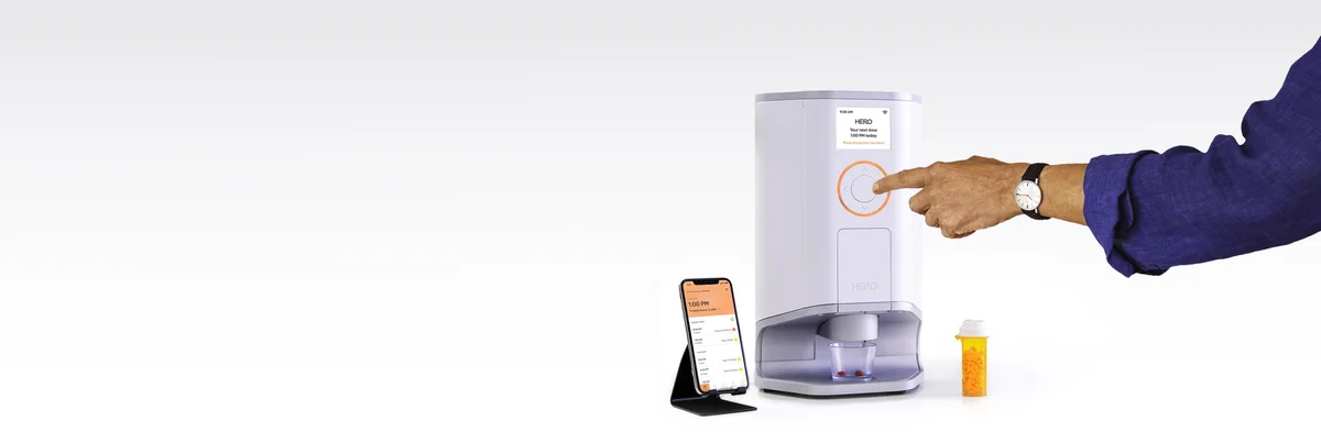 Hero Smart Pill Dispenser on a countertop being operated by hand, with companion app on a phone and a pill bottle alongside