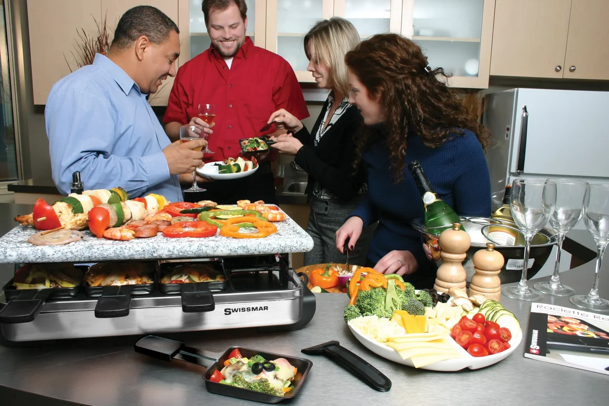 Friends gathered around the Swissmar Stelvio granite stone raclette grill at a kitchen party, with skewers and peppers cooking on the stone