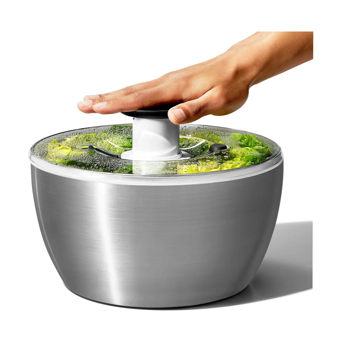 Hand pressing the black pump knob of the OXO Steel Salad Spinner to spin wet lettuce inside the stainless steel bowl