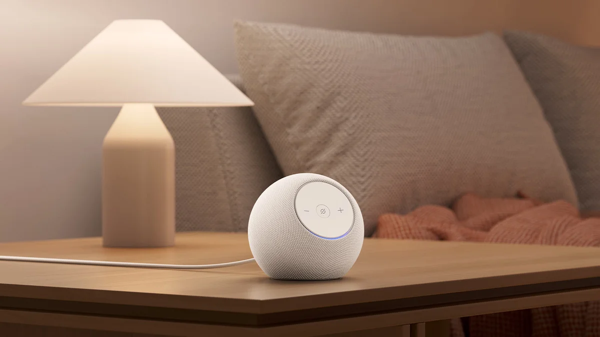 Amazon Echo Dot Max Glacier White on a bedside table beside a lamp, in a cozy bedroom setting
