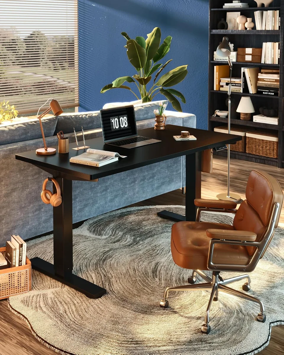 Black FlexiSpot EN1 standing desk in a living room beside a sofa and bookshelf, with a laptop, desk lamp and a leather office chair, headphones hanging from the side hook