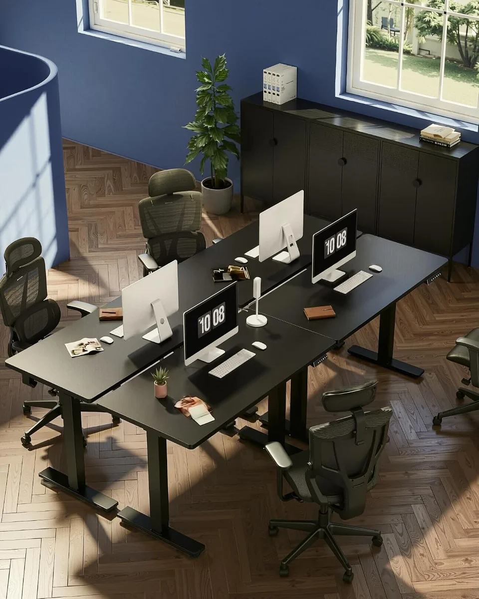 Overhead view of several black FlexiSpot EN1 standing desks set up in pairs in a shared office with mesh chairs and monitors, showing the T-leg frames and one-piece desktops