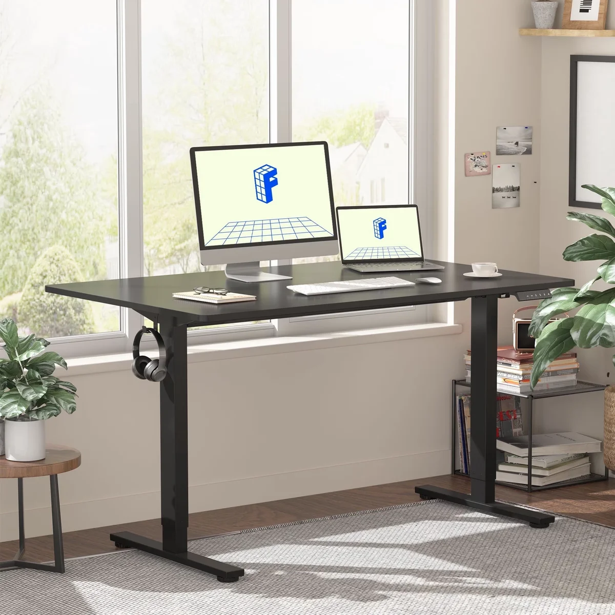 FlexiSpot EN1 electric standing desk with black one-piece top and black single-motor T-leg frame in a bright home office by a window, showing the small keypad at the front right edge and a headphone hook on the left leg