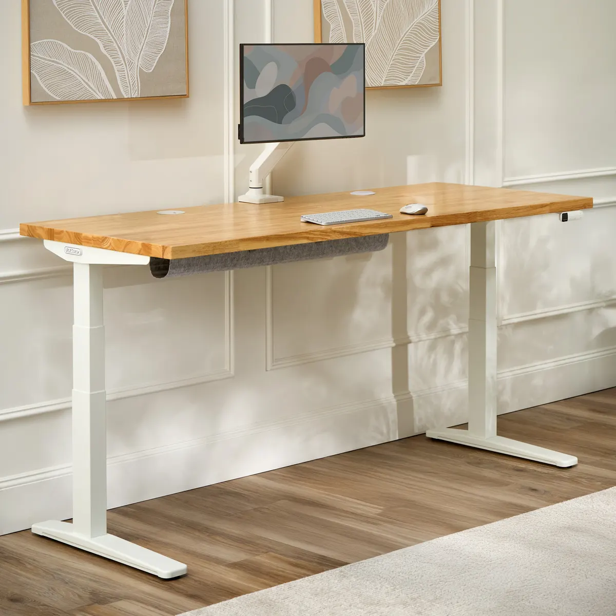 UPLIFT V3 Standing Desk with a natural solid wood top and white 3-stage frame with C-frame feet, full desk view with a monitor arm and felt cable sleeve against a paneled wall