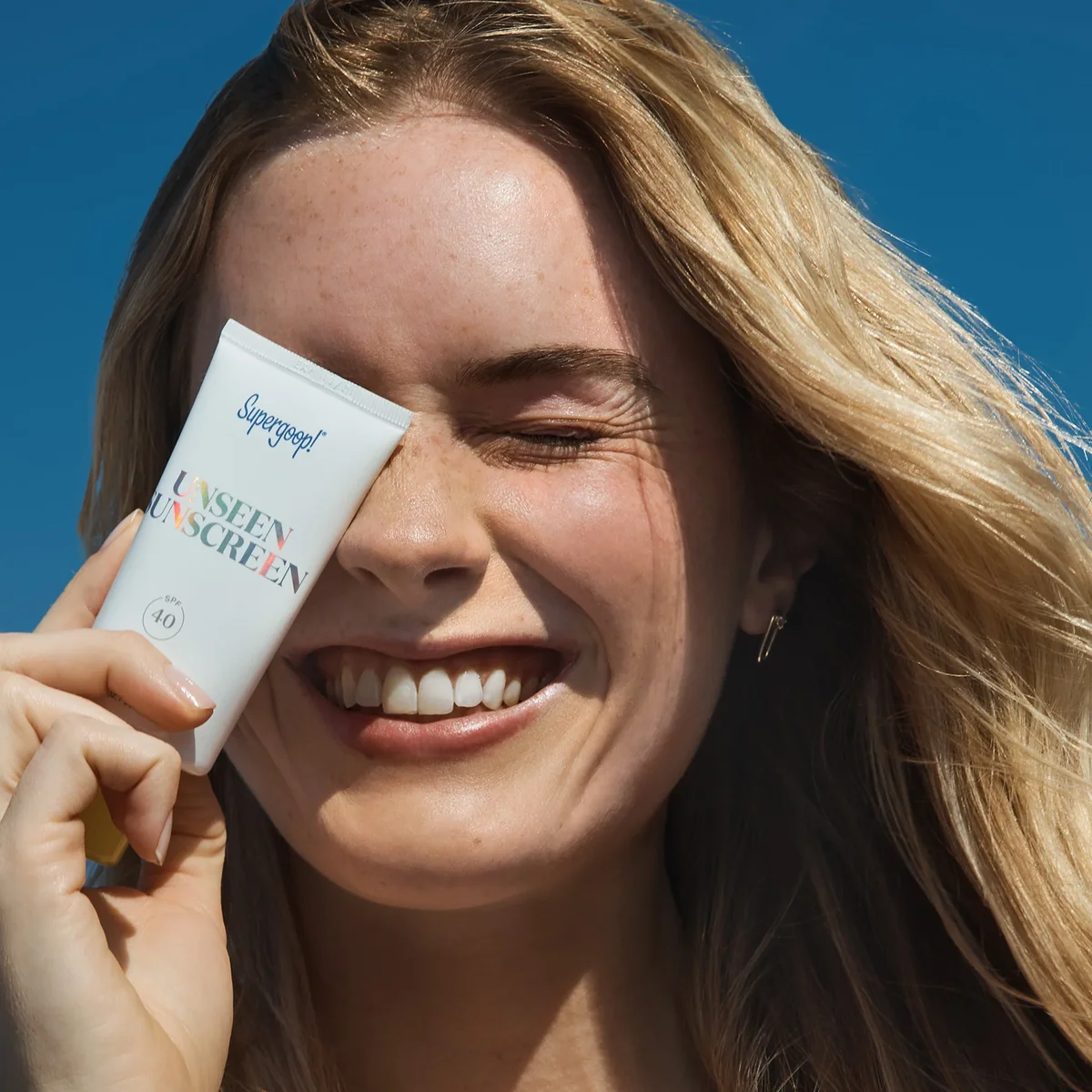 Woman holding Supergoop! Unseen Sunscreen SPF 40 tube outdoors against blue sky