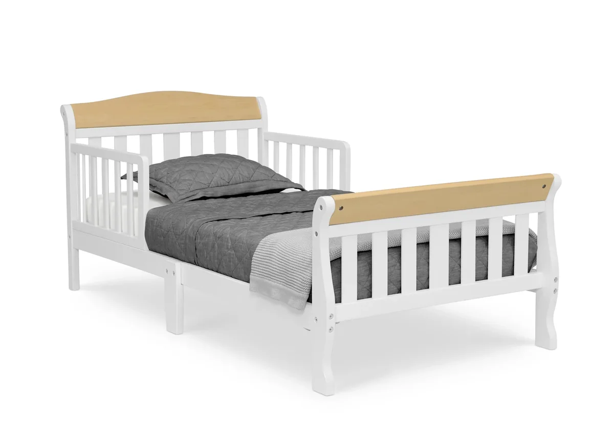 Delta Children Canton Toddler Bed in white with natural wood accents, 3/4 angle showing both attached side guardrails