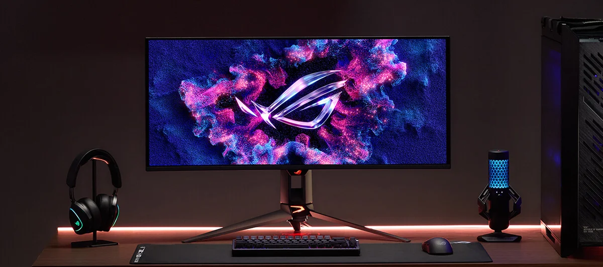 ASUS ROG Swift OLED PG34WCDN gaming desk setup with headset, keyboard, and microphone in ambient lighting