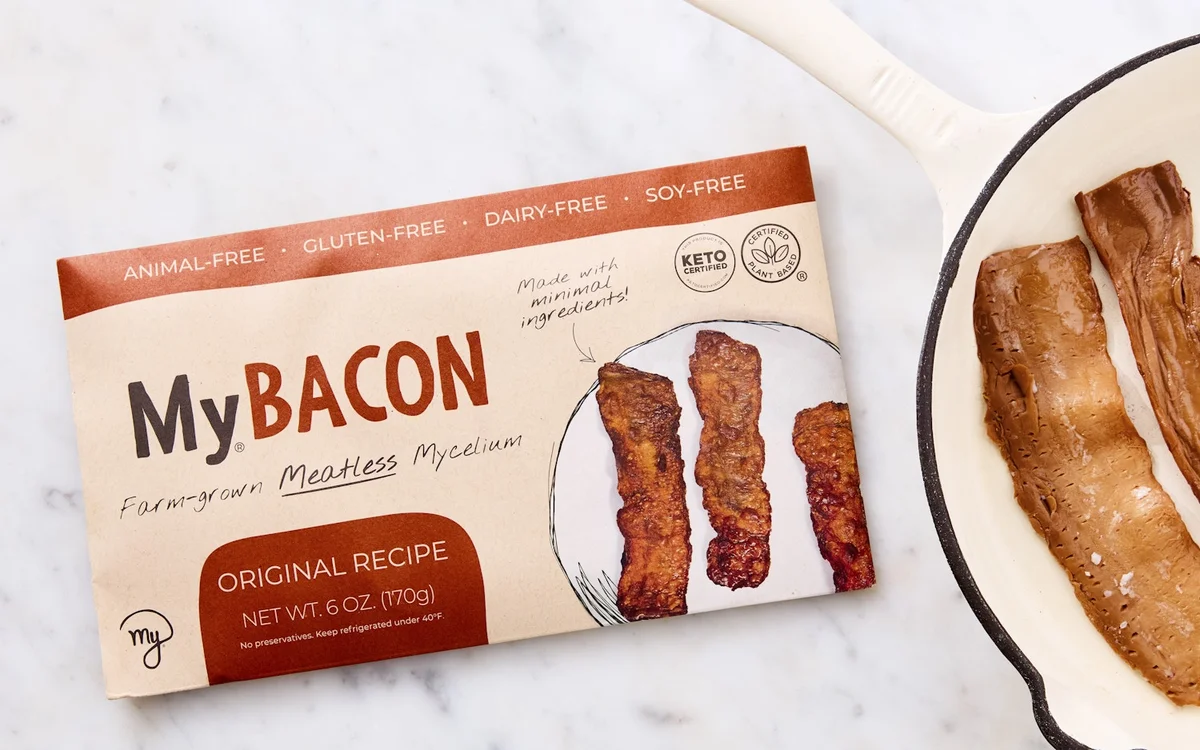MyBacon Original Recipe package next to cooked mycelium bacon strips in a cast iron skillet on a marble surface