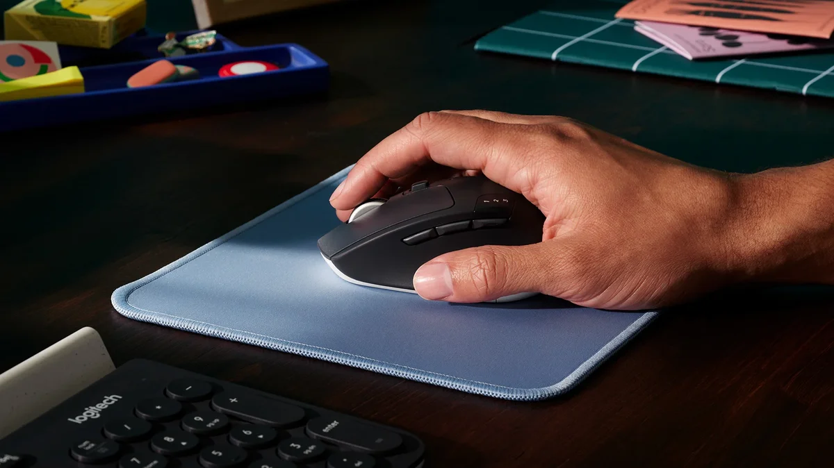 Hand gripping the Logitech M720 Triathlon on a blue mouse pad at a desk, thumb resting beside the Easy-Switch 1-2-3 button and thumb buttons